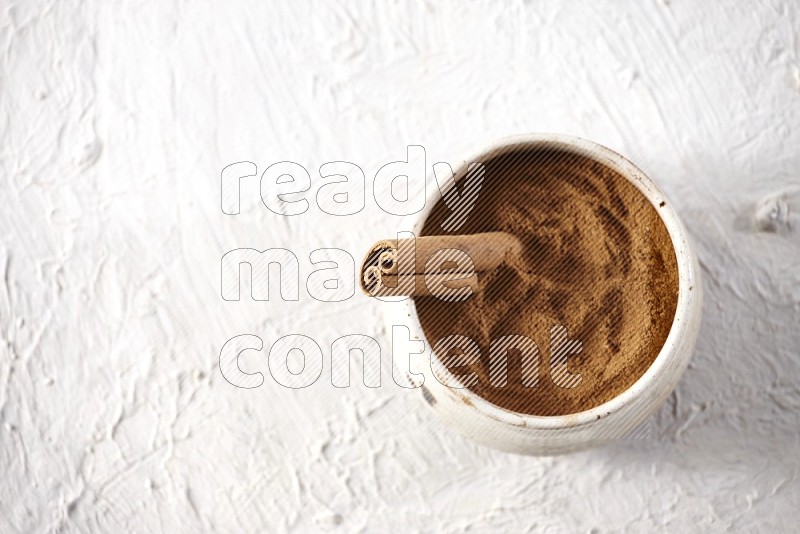 Ceramic beige bowl full of cinnamon powder with a cinnamon stick on a textured white background