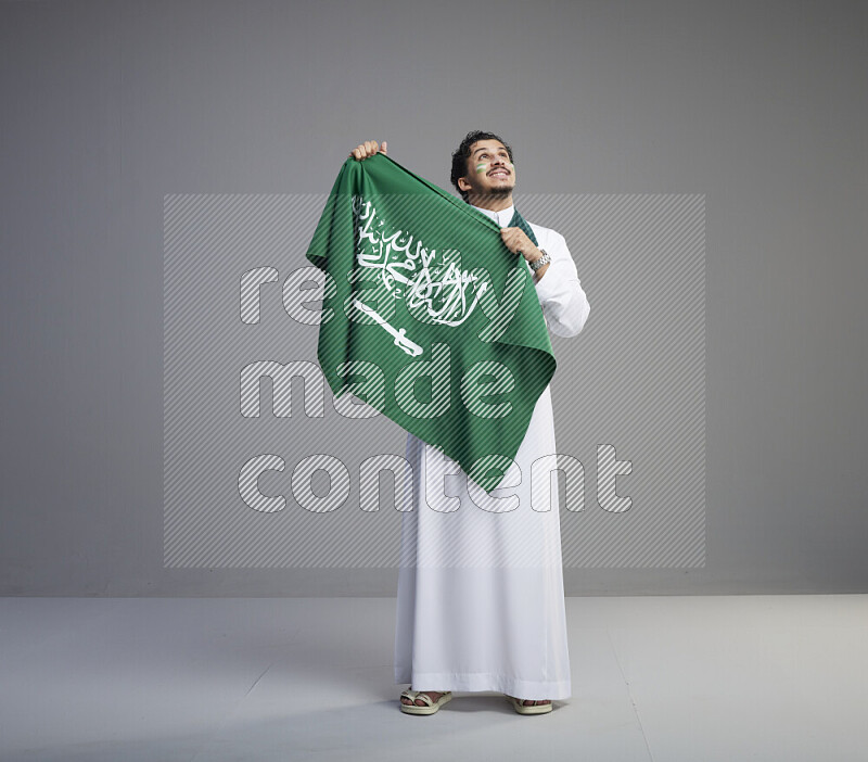 A Saudi man standing wearing thob with face painting wrapping Saudi flag scarf and holding big Saudi flag on gray background