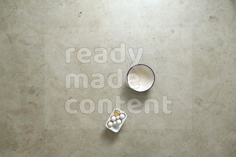 A ceramic bowl beside a carton of eggs and glass bowl full of sugar, vanilla extract, flour on beige marble background