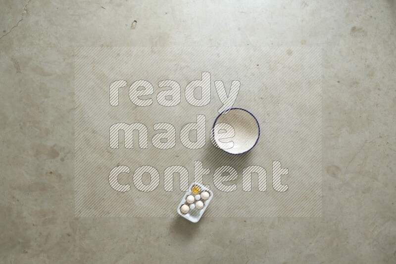 A ceramic bowl beside a carton of eggs and glass bowl full of sugar, vanilla extract, flour on beige marble background
