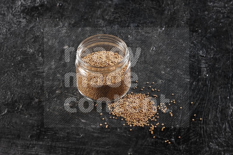 A glass jar full of mustard seeds and jar is flipped and seeds spread out on a textured black flooring in different angles