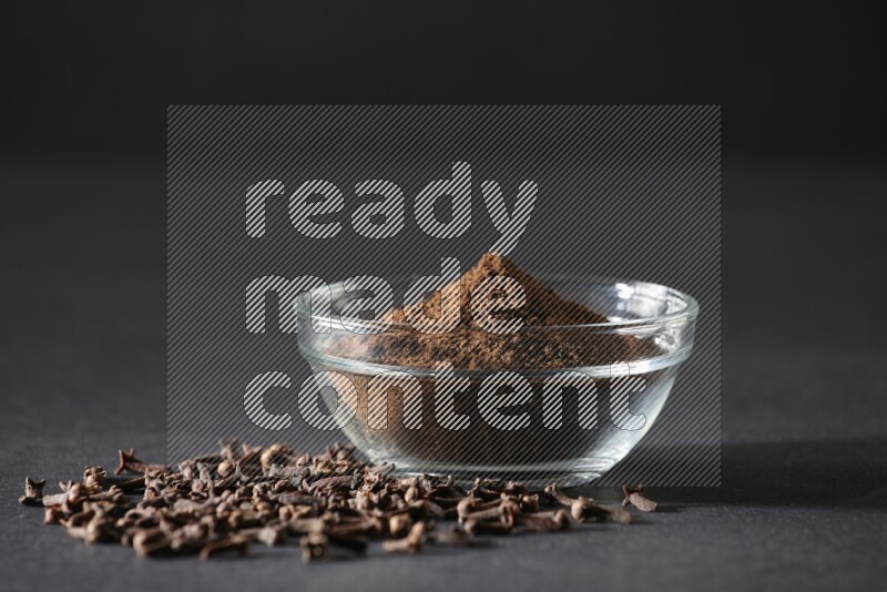 A glass bowl full of cloves powder with cloves grains spread on black flooring