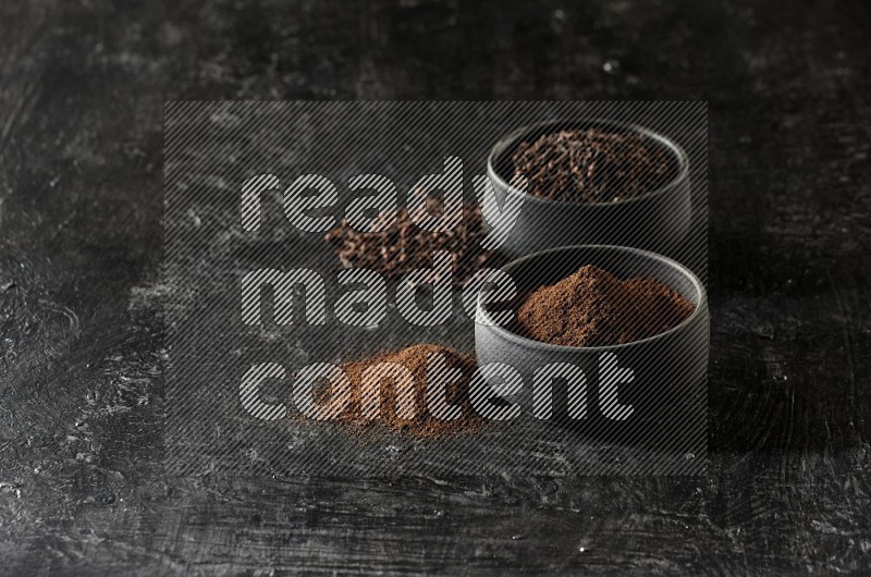 2 Black pottery bowls full of cloves and the other full of cloves powder on textured black flooring