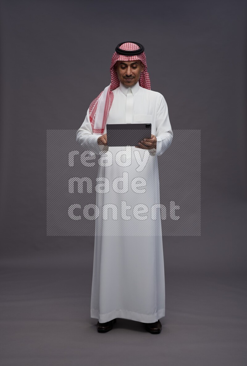 Saudi man wearing thob and shomag standing working on tablet on gray background