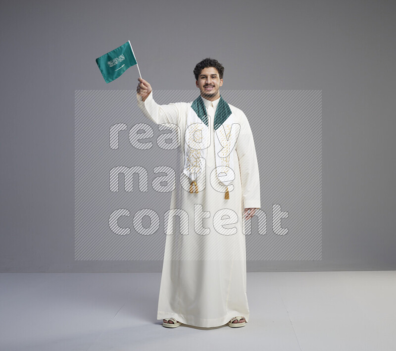 A Saudi man standing wearing thob and saudi flag scarf and holding small saudi flag on gray background