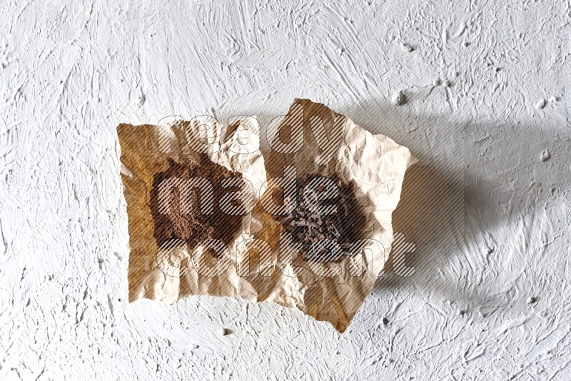 Cloves powder and cloves on 2 crumpled pieces of paper on a textured white flooring