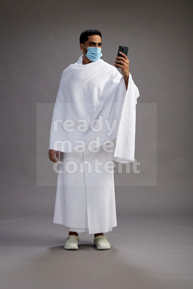 A man wearing Ehram with face mask Standing taking selfie on gray background