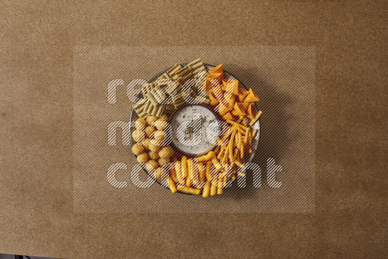 Assorted snacks on a pottery plate with a dipping on brown background