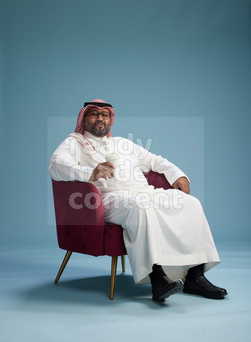 Saudi Man with shimag sitting on chair drinking coffee on blue background