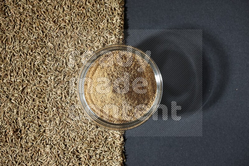 A glass bowl full of cumin powder and cumin seeds beneath it on black flooring