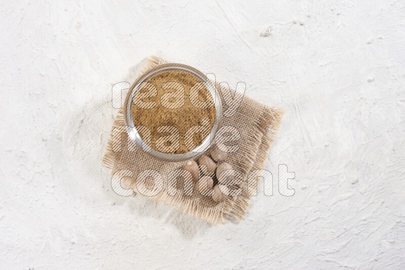 A glass bowl full of nutmeg powder with whole seeds beside it on burlap fabric on a textured white flooring