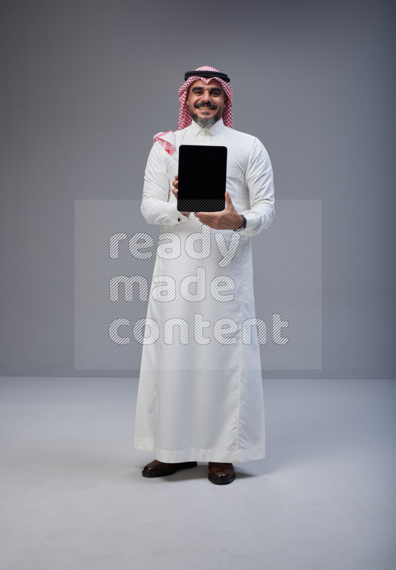 Saudi man Wearing Thob and red Shomag standing showing tablet to camera on Gray background
