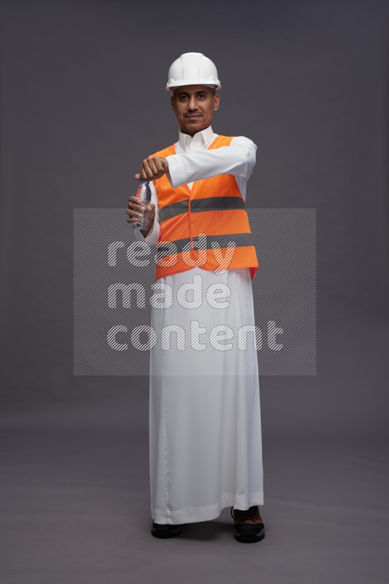 Saudi man wearing thob with engineer vest standing drinking water on gray background