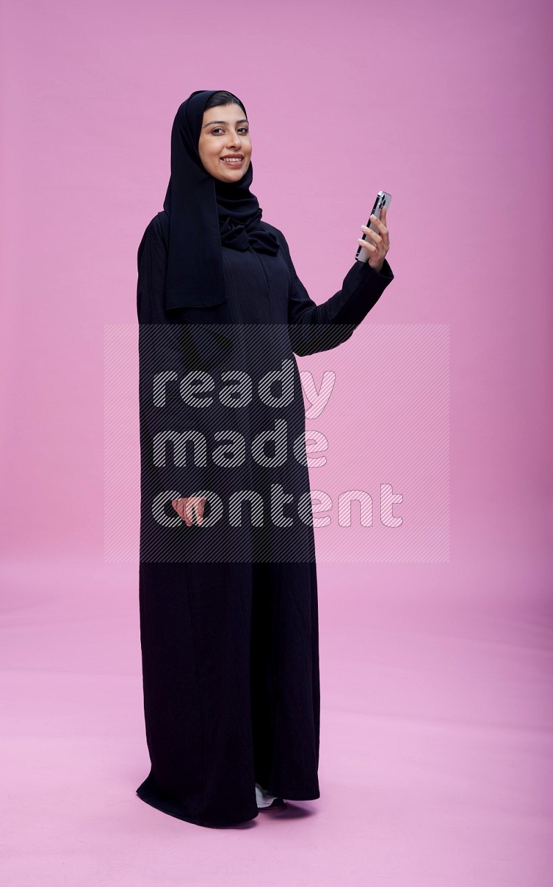 Saudi woman wearing Abaya standing taking selfie on pink background