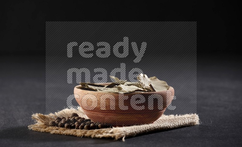 A wooden bowl filled with dried bay leaves on a piece of burlap with bunch of allspice berries on black flooring