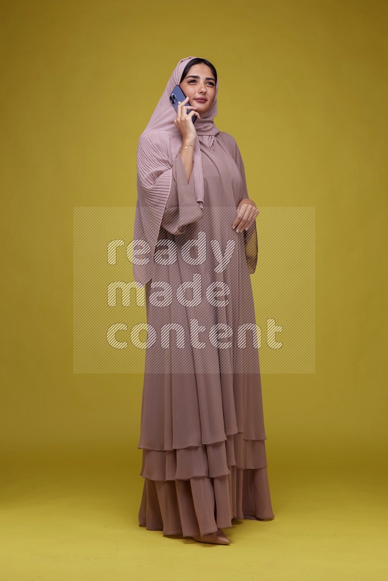 A woman having a call on a Yellow Background wearing Brown Abaya with Hijab