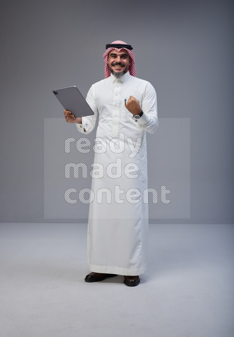 Saudi man Wearing Thob and red Shomag standing working on tablet on Gray background