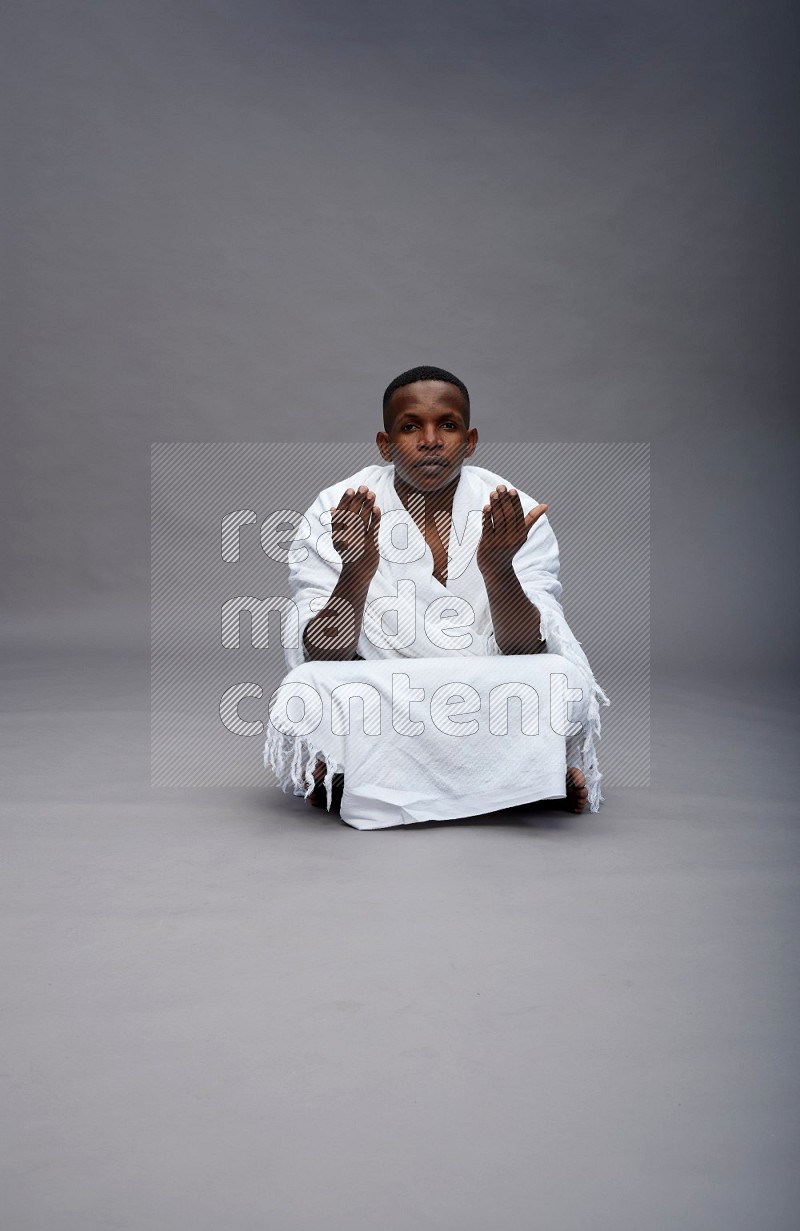 A man wearing Ehram sitting on floor dua'a on gray background