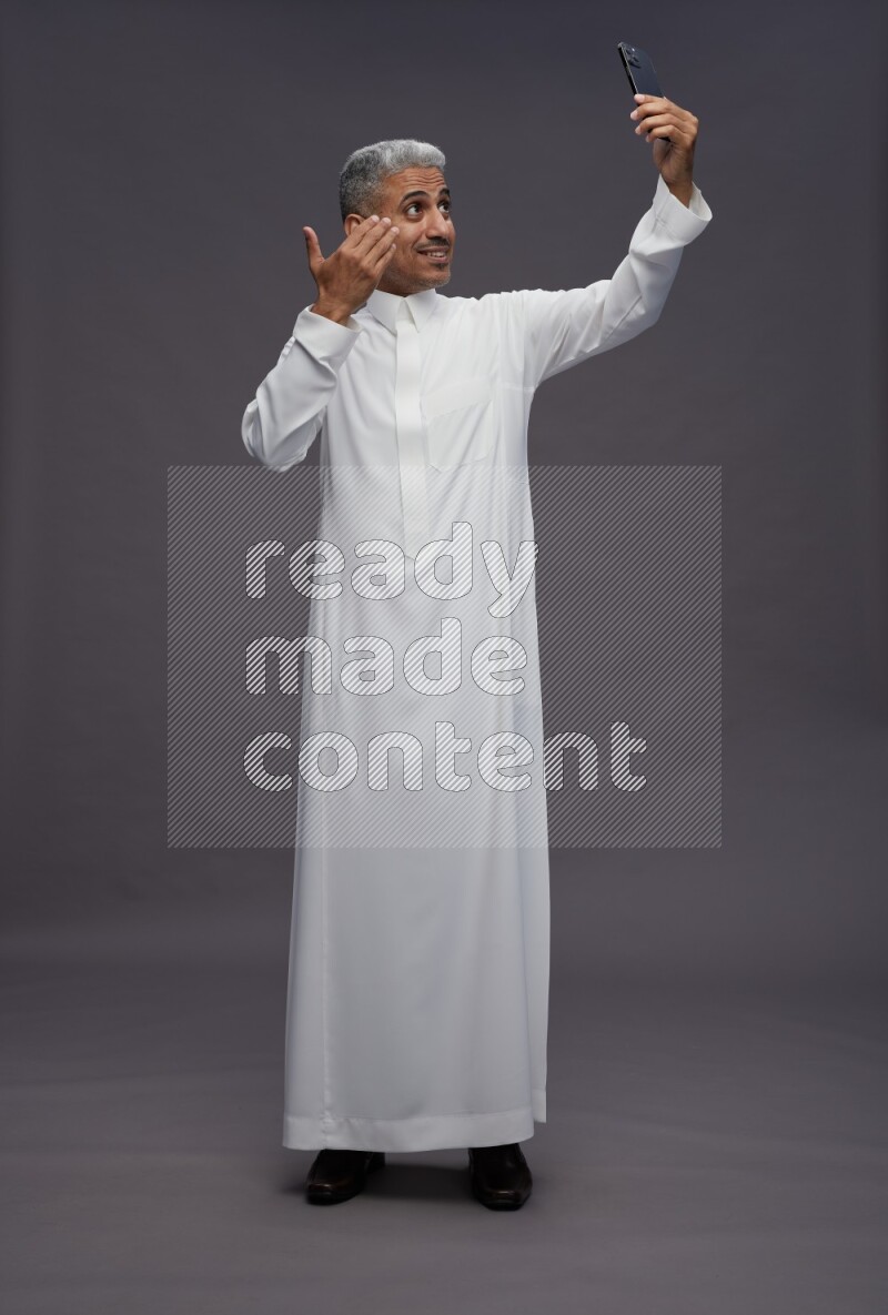 Saudi man wearing thob standing taking selfie on gray background