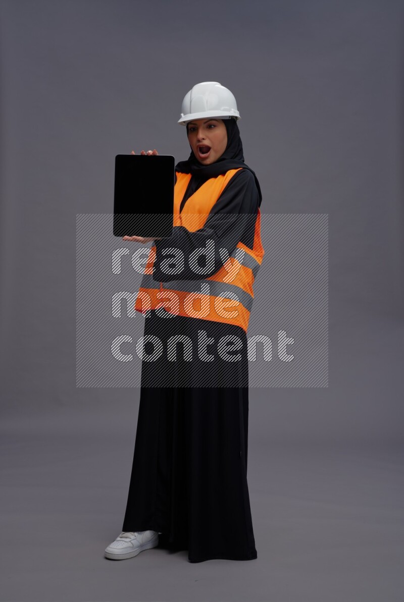 Saudi woman wearing Abaya with engineer vest standing showing tablet to camera on gray background