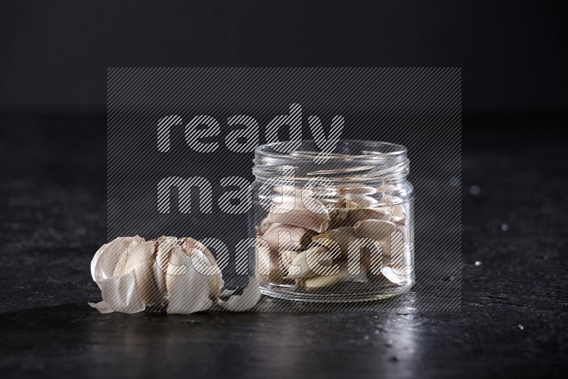 A glass jar full of garlic cloves and beside it a garlic bulb on a textured black flooring in different angles