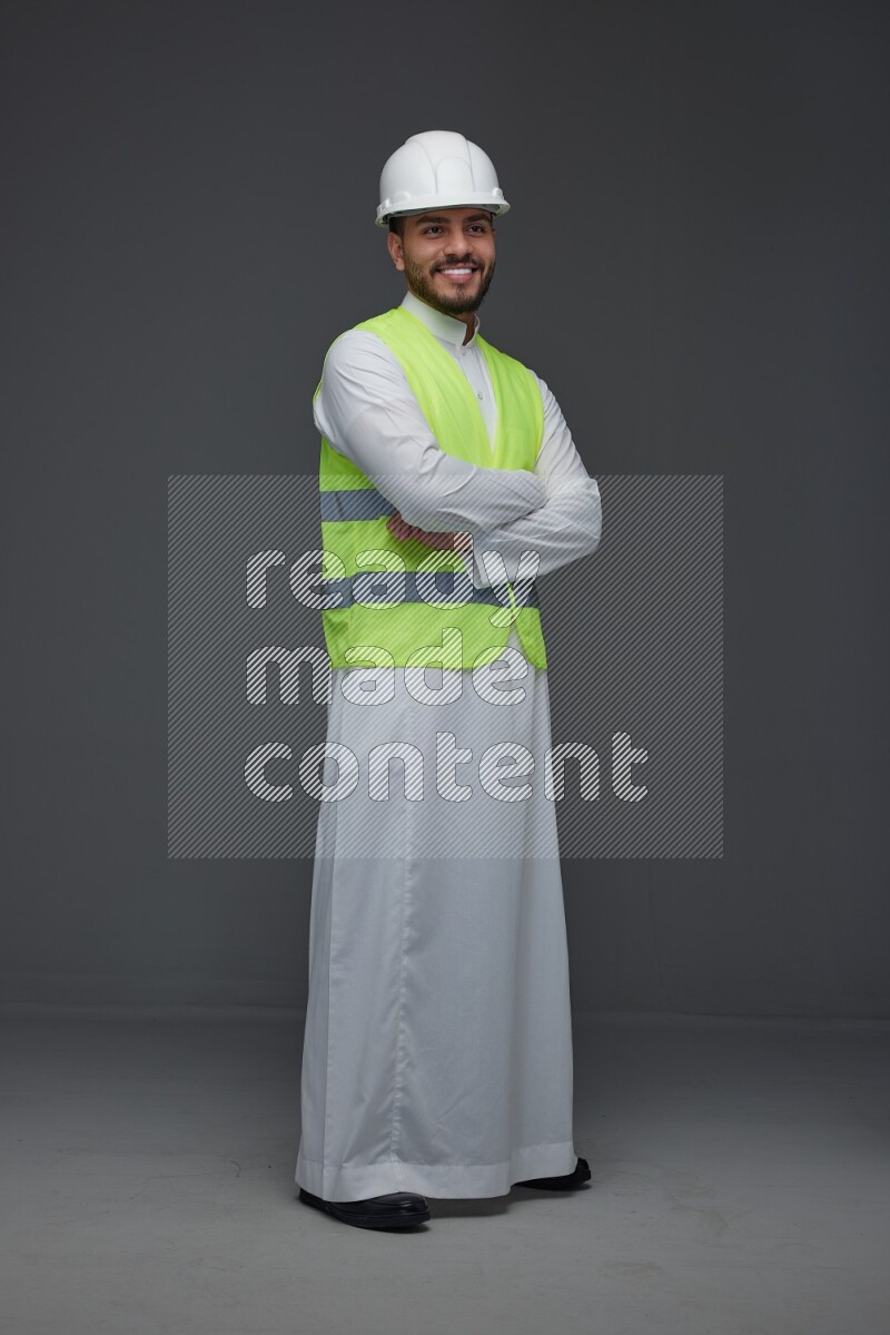 A Saudi man wearing Thobe with a yellow safety vest and white helmet standing and crossing his hands eye level on a gray background