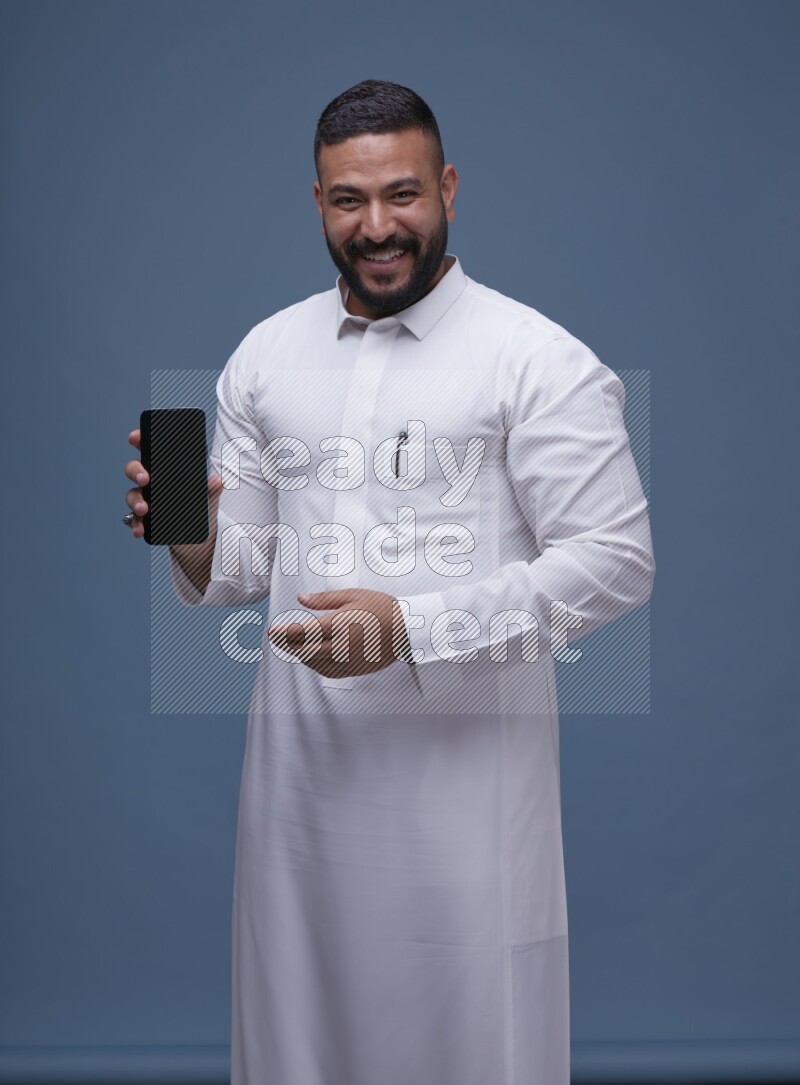 A man Showing his phone in Blue Background wearing Saudi Thob