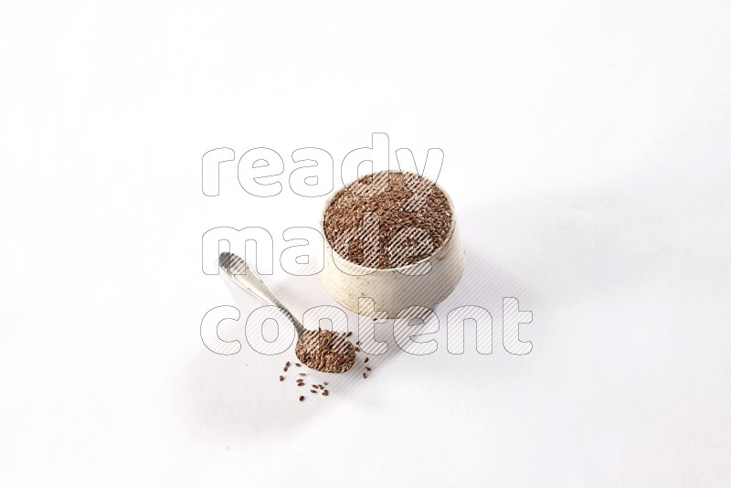 A pottery beige bowl and a metal spoon full of flax seeds on a white flooring