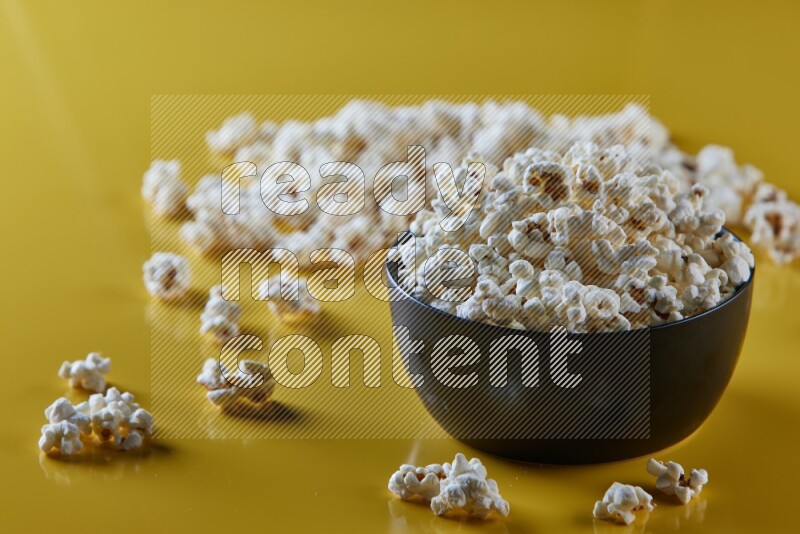 A black ceramic bowl full of popcorn with popcorn beside it on a yellow background in different angles