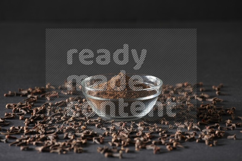 A glass bowl full of cloves powder with cloves grains spread on black flooring