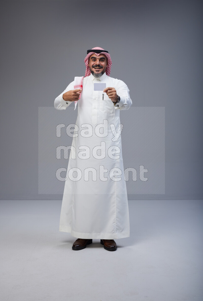 Saudi man Wearing Thob and red Shomag standing holding ATM card on Gray background