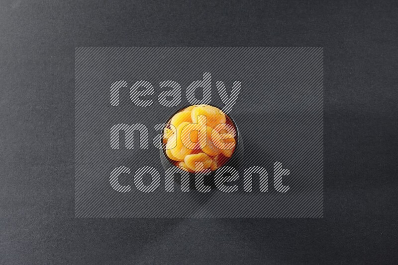 A black pottery bowl full of dried apricots on a black background in different angles