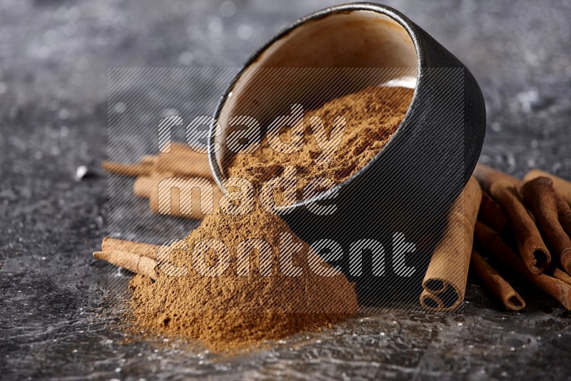 Black pottery bowl over filled with cinnamon powder and cinnamon sticks around the bowl on a textured black background