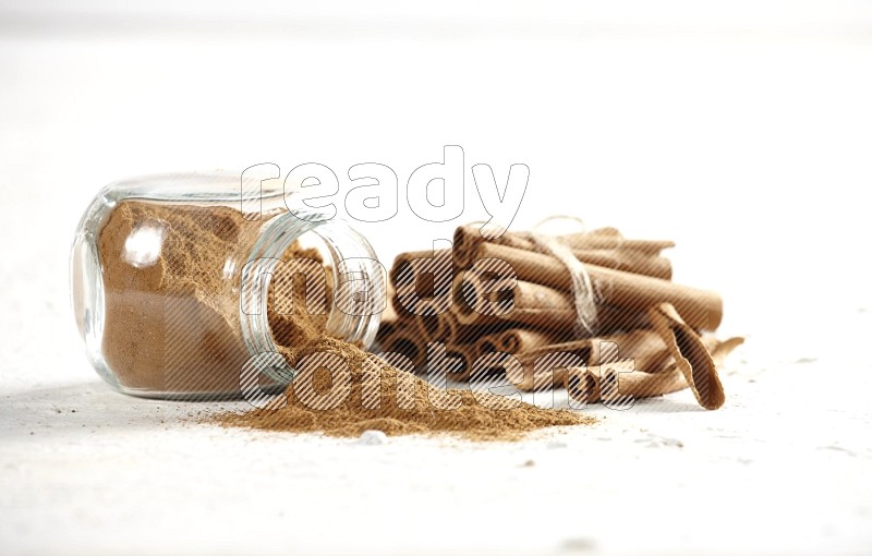 Flipped herbs glass jar full of cinnamon powder and cinnamon sticks in the back on a textured white background