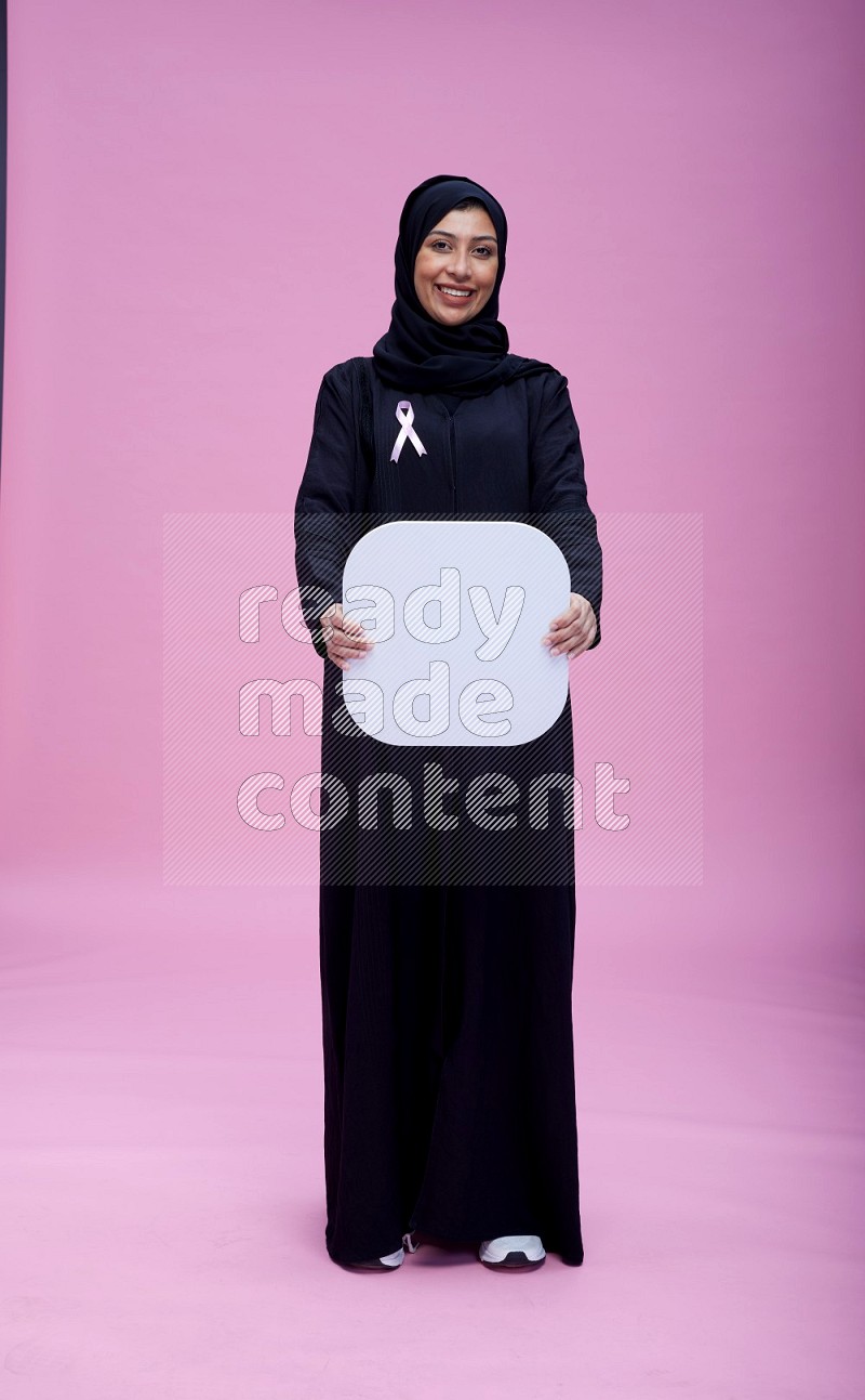 Saudi woman wearing pink ribbon on Abaya standing holding social media on pink background
