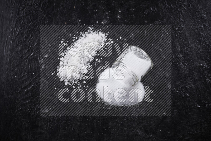 A glass jar full of table salt with some sea salt crystals beside it on a black background