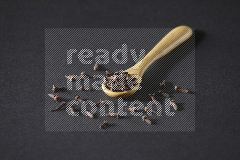 A wooden spoon full of cloves and some spreaded out of it on a black flooring