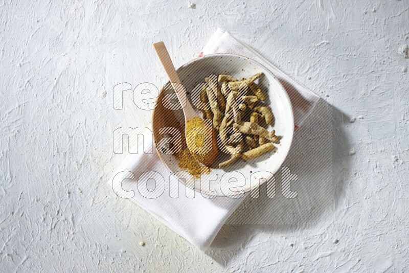 A plate filled with dried turmeric whole fingers and a wooden spoon full of turmeric powder on a textured white flooring