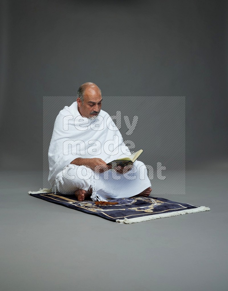 A man wearing Ehram sitting on mate prayer reading quran on gray background