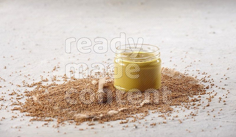 A glass jar full of mustard paste set on a burlap piece with a wooden spoon full of mustard seeds on a textured white flooring