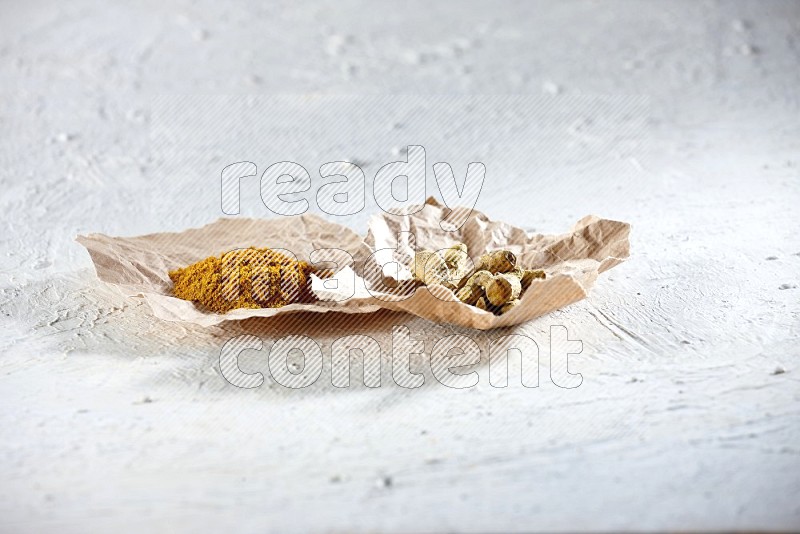 Turmeric powder and dried turmeric whole fingers in 2 crumpled pieces of paper on textured white flooring
