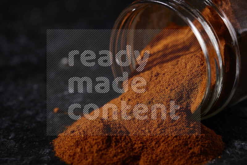 A glass jar full of ground paprika powder flipped with some spilling powder on black background