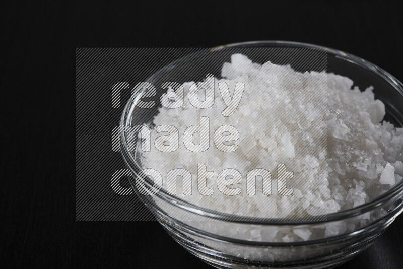 A glass bowl full of coarse sea salt crystals on black background