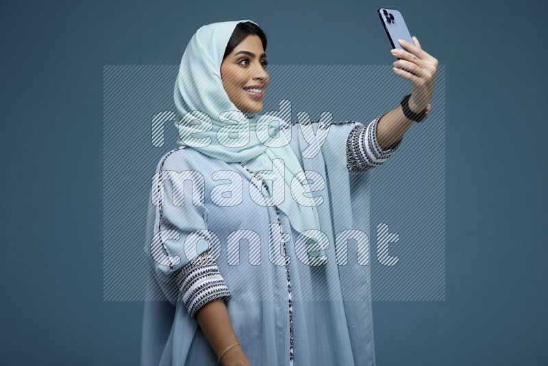 A Saudi woman Taking a Selfie on a blue background wearing a blue Abaya with hijab
