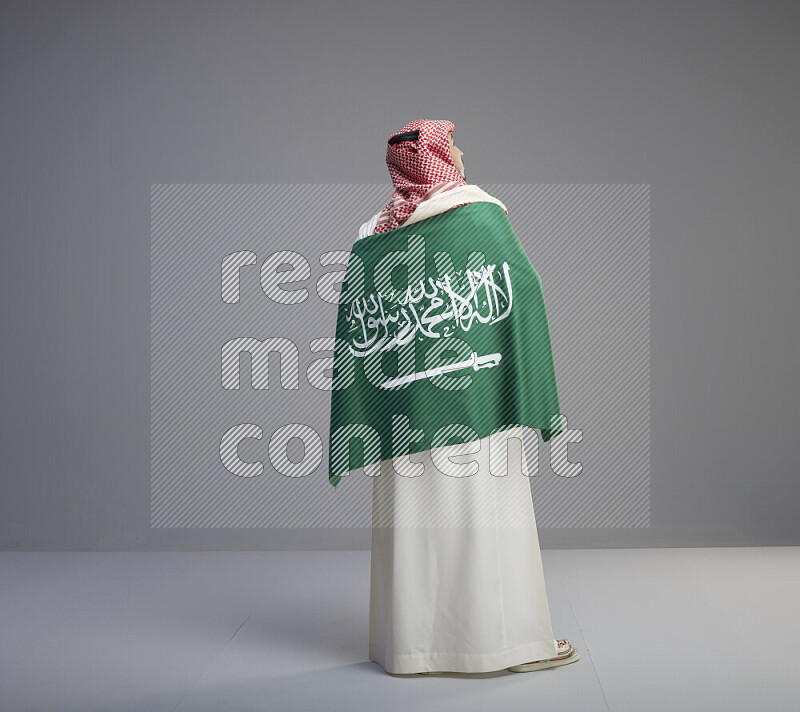 رجل سعودي يرتدي ثوب ابيض وشماغ احمر يحمل العلم السعودي على كتفيه