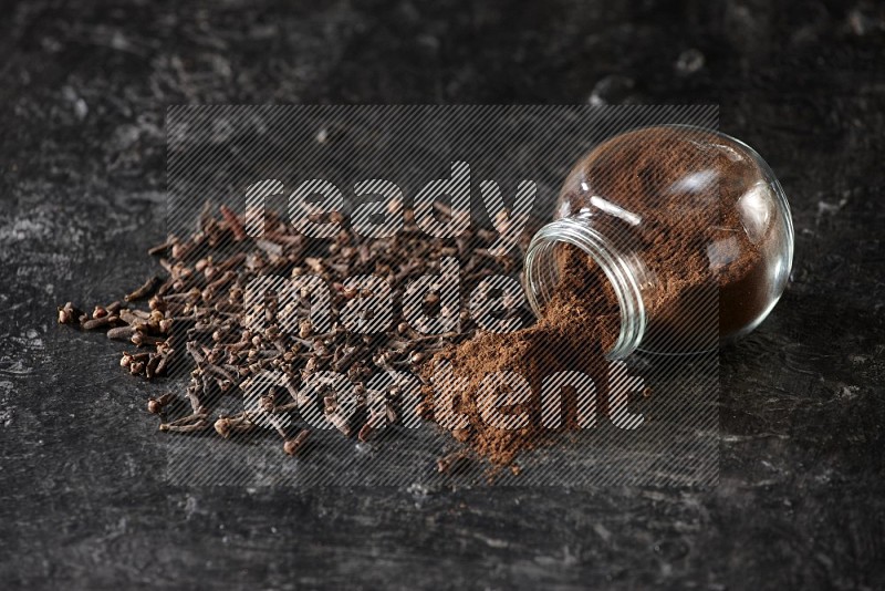 A flipped glass spice jar full of cloves powder and powder came out of it with cloves spread on textured black flooring