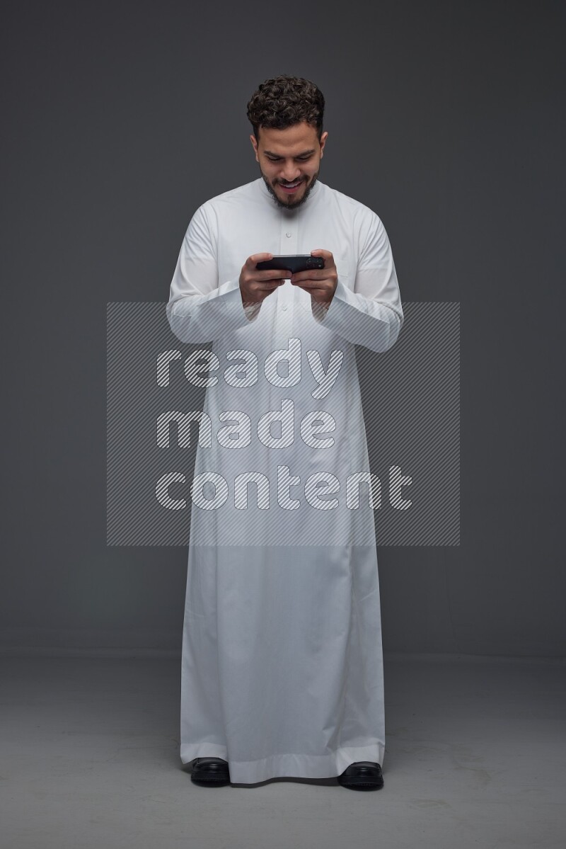 A Saudi man wearing Thobe and playing using his phone horizontally playing video games while standing and making different poses eye level on a gray background