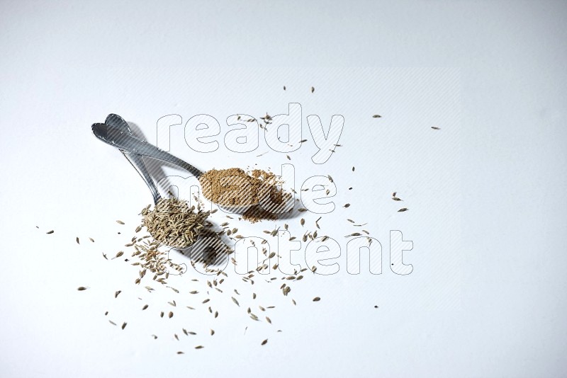 2 metal spoons full of cumin seeds and powder on white flooring