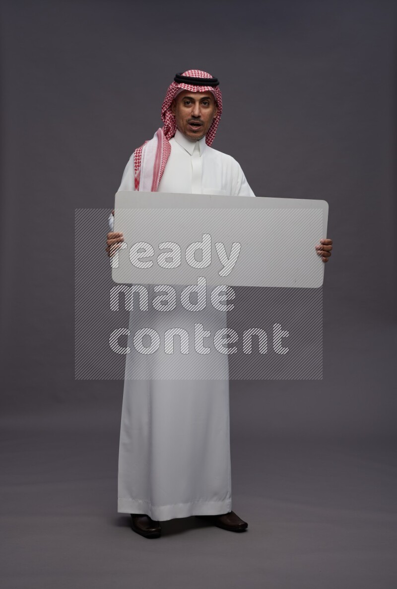 Saudi man wearing thob and shomag standing holding board on gray background