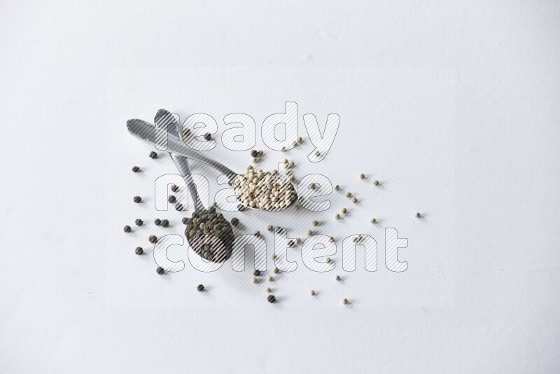 ملعقتان معدنيتان ممتلئتان بحبوب الفلفل الأسود والأبيض مع حبوب متناثرة على أرضية بيضاء